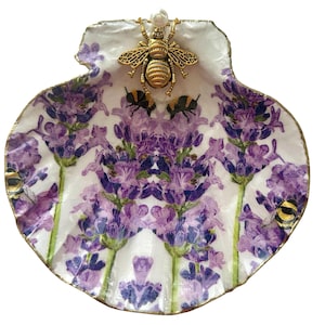 May include: A decorative seashell dish featuring a lavender floral design with a gold rim. The dish is adorned with a gold-colored bee and several smaller bee accents. The background is white.