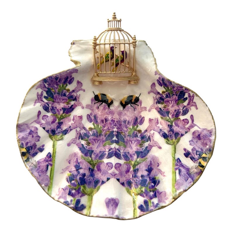 May include: A decorative scallop shell dish featuring a lavender floral pattern with two bumblebees and a small gold-colored birdcage ornament attached to the top edge, containing two tiny birds inside the cage structure.