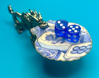 May include: A brass dragon figurine attached to a scalloped, shell-shaped dish featuring a blue and white pattern with a dragon illustration. Two translucent blue dice with white pips are resting inside the dish, showing three and five.