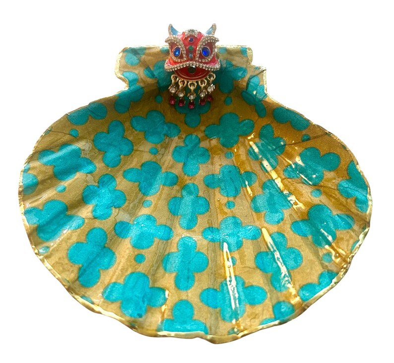 Puede incluir: Un plato decorativo en forma de concha con un patr&oacute;n floral dorado y turquesa. El plato tiene un adorno de joyas en rojo, azul y dorado. El adorno tiene cuentas rojas colgantes. Un art&iacute;culo de decoraci&oacute;n del hogar &uacute;nico.