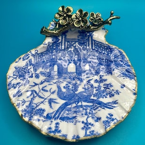 May include: A decorative scallop shell dish featuring a blue and white chinoiserie pattern of a bridge and two birds. The top edge is adorned with a metallic brass-toned branch with three flowers. The rim of the shell is finished in gold paint.