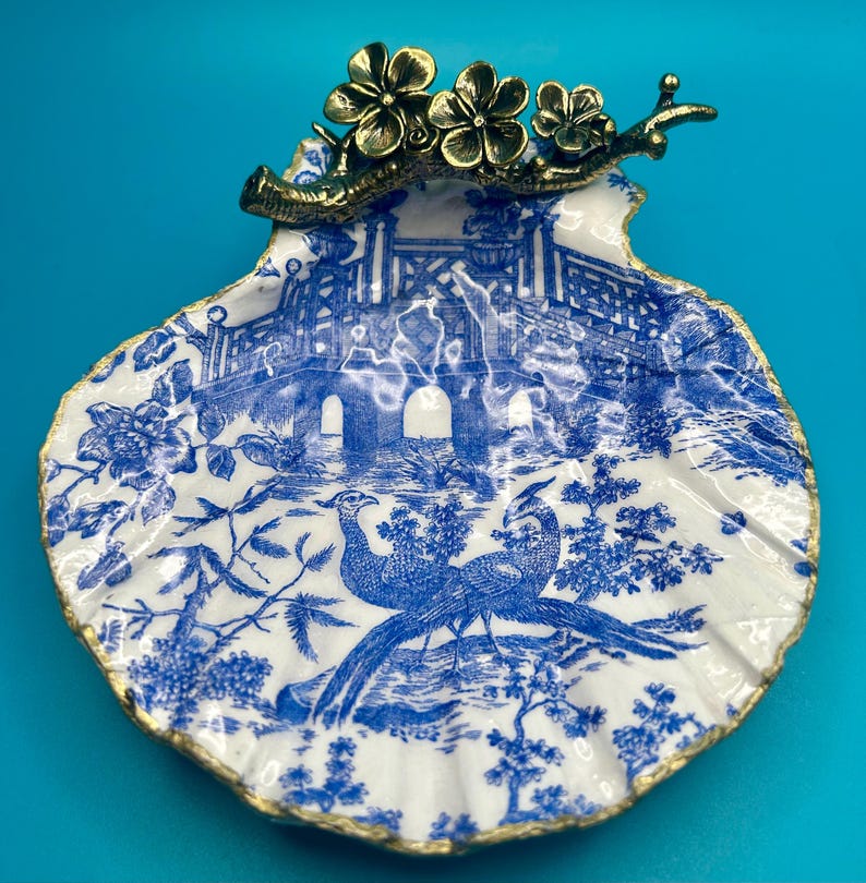 May include: A decorative scallop shell dish featuring a blue and white chinoiserie pattern of a bridge and two birds. The top edge is adorned with a metallic brass-toned branch with three flowers. The rim of the shell is finished in gold paint.