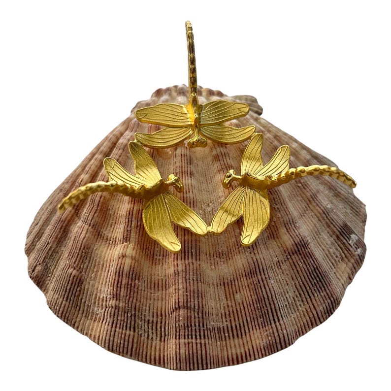 May include: Three gold-colored dragonfly ornaments arranged in a circular formation. Each dragonfly features detailed wings with linear textures and a segmented body. The ornaments are positioned on top of a large, fan-shaped, brown-striped seashell.