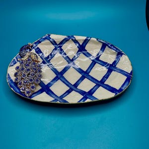 May include: A decorative, oval-shaped ceramic tray with a white base and a blue grid pattern. A cluster of blue gemstones adorns the edge. The tray is set against a solid blue background.
