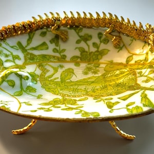 May include: An oval-shaped decorative dish with a white and green floral design. The dish is supported by three gold-colored legs and is adorned with a gold-colored dragon along the rim. The dragon has red accents on its head.