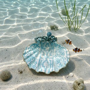 May include: An ornate seashell with a blue and white geometric design, topped with a beaded crown and a blue jewel. The underwater scene includes two clownfish. The shell sits on a sandy seabed, with a few plants.