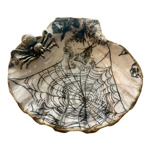 May include: A decorative seashell-shaped dish with a Halloween theme. The dish features a black spiderweb design and a bronze-colored spider attached to the side. The edges are trimmed with gold.