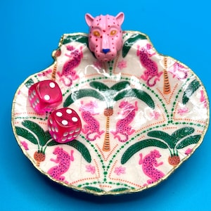 Op de afbeelding: Een decoratieve schelpvormige schaal met een handgeschilderd patroon van roze luipaarden en groene palmbomen op een cr&egrave;mekleurige achtergrond. Een gebeeldhouwde roze luipaardkop is aan de bovenrand bevestigd. Twee roze dobbelstenen liggen erin.