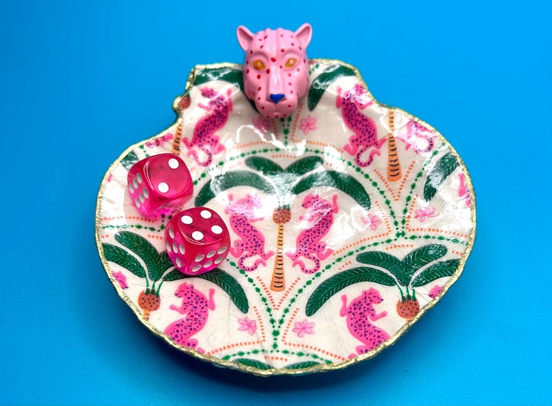 Op de afbeelding: Een decoratieve schelpvormige schaal met een handgeschilderd patroon van roze luipaarden en groene palmbomen op een cr&egrave;mekleurige achtergrond. Een gebeeldhouwde roze luipaardkop is aan de bovenrand bevestigd. Twee roze dobbelstenen liggen erin.
