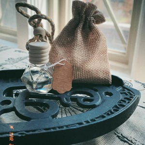 May include: A clear, faceted glass bottle with a wooden cap and a hanging rope, next to a small burlap bag and a brown tag. The items are on a black, decorative metal tray. The date "MAR 28 '25" is visible on the tray.