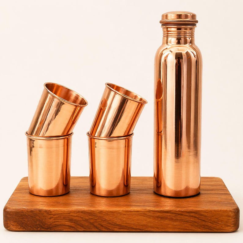 May include: A copper water bottle with a screw-on cap and three copper cups are displayed on a wooden board. The bottle has a slender design with a small handle. The cups are cylindrical with a slight outward flare at the top.