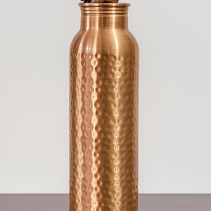 May include: A copper-colored water bottle with a textured, hammered finish. The bottle has a screw-on lid that is partially unscrewed. The bottle is cylindrical and has a capacity of approximately 25 ounces.