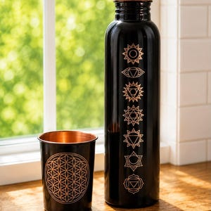 K&ouml;nnte beinhalten: Eine schwarze Metallwasserflasche mit kupferfarbenem Verschluss und sieben vertikalen Chakrasymbolen in Kupfer. Daneben steht ein passender schwarzer Metallbecher mit kupferfarbenem Inneren und einem geometrischen Blume-des-Lebens-Muster in Kupfer.