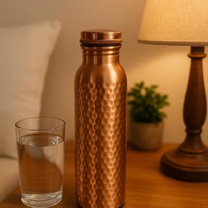May include: A copper water bottle with a textured surface and a matching cap. A glass of water sits next to the bottle on a wooden surface. A lamp and small plant are in the background.
