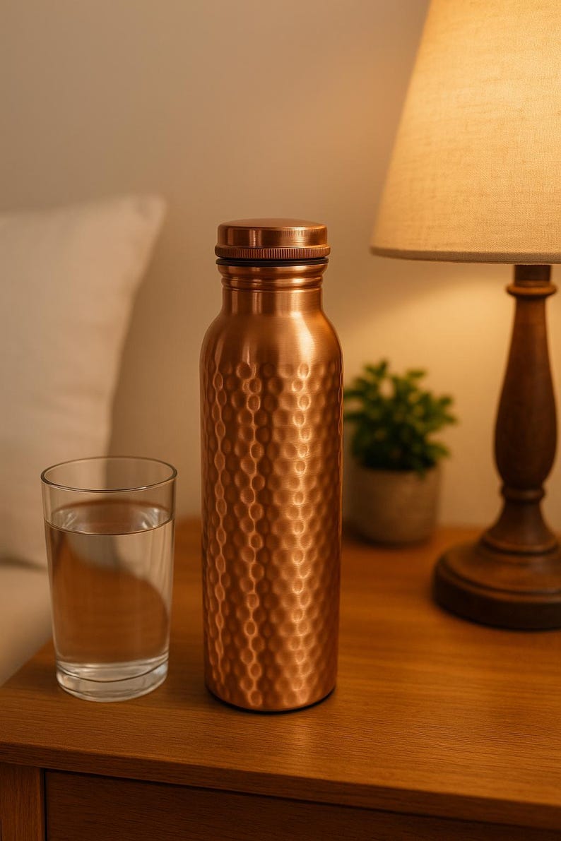 May include: A copper water bottle with a textured surface and a matching cap. A glass of water sits next to the bottle on a wooden surface. A lamp and small plant are in the background.