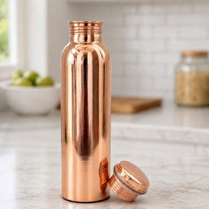 May include: A copper-colored water bottle with a matching screw-on lid. The bottle is cylindrical with a polished finish, reflecting the surrounding environment. The lid is lying next to the bottle. The bottle is on a marble countertop.
