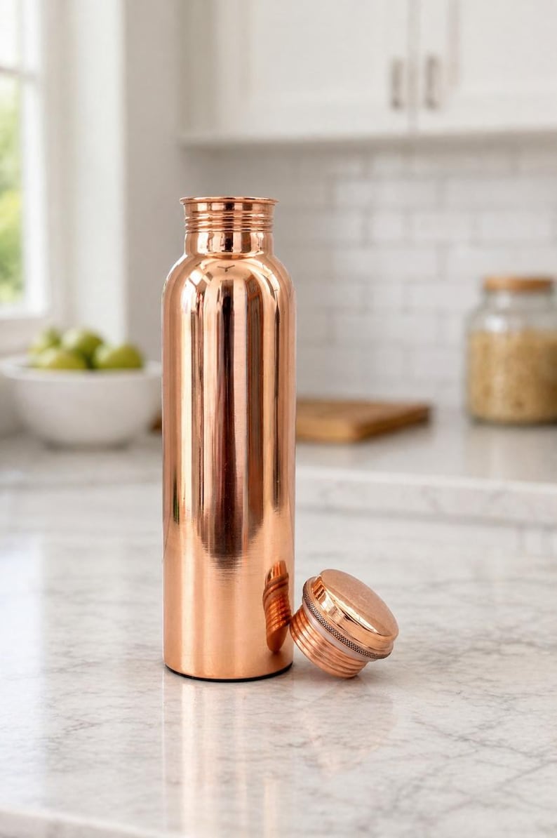 May include: A copper-colored water bottle with a matching screw-on lid. The bottle is cylindrical with a polished finish, reflecting the surrounding environment. The lid is lying next to the bottle. The bottle is on a marble countertop.
