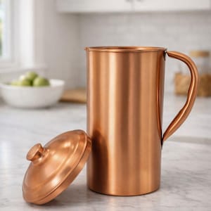 May include: A copper pitcher with a matching lid. The pitcher has a curved handle and a cylindrical body. The lid is round with a small knob. The pitcher and lid are sitting on a white countertop. The background is a kitchen.