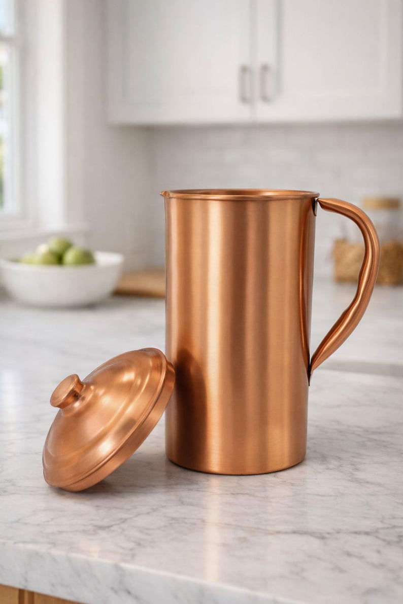 May include: A copper pitcher with a matching lid. The pitcher has a curved handle and a cylindrical body. The lid is round with a small knob. The pitcher and lid are sitting on a white countertop. The background is a kitchen.
