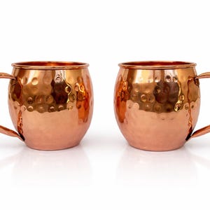 May include: Two copper Moscow Mule mugs with a hammered finish. Each mug has a rounded body, a curved handle, and a shiny, reflective surface. The mugs are displayed on a white background.