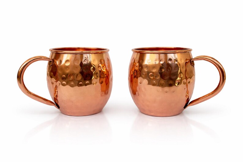 May include: Two copper Moscow Mule mugs with a hammered finish. Each mug has a rounded body, a curved handle, and a shiny, reflective surface. The mugs are displayed on a white background.