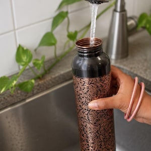 K&ouml;nnte beinhalten: Eine zylindrische Wasserflasche aus Kupfer mit schwarzem Finish an Hals und Boden sowie einem detaillierten, ge&auml;tzten Blumenmuster in metallischem Kupfer auf dem Hauptk&ouml;rper. Die Flasche wird gerade unter einem Wasserhahn bef&uuml;llt.