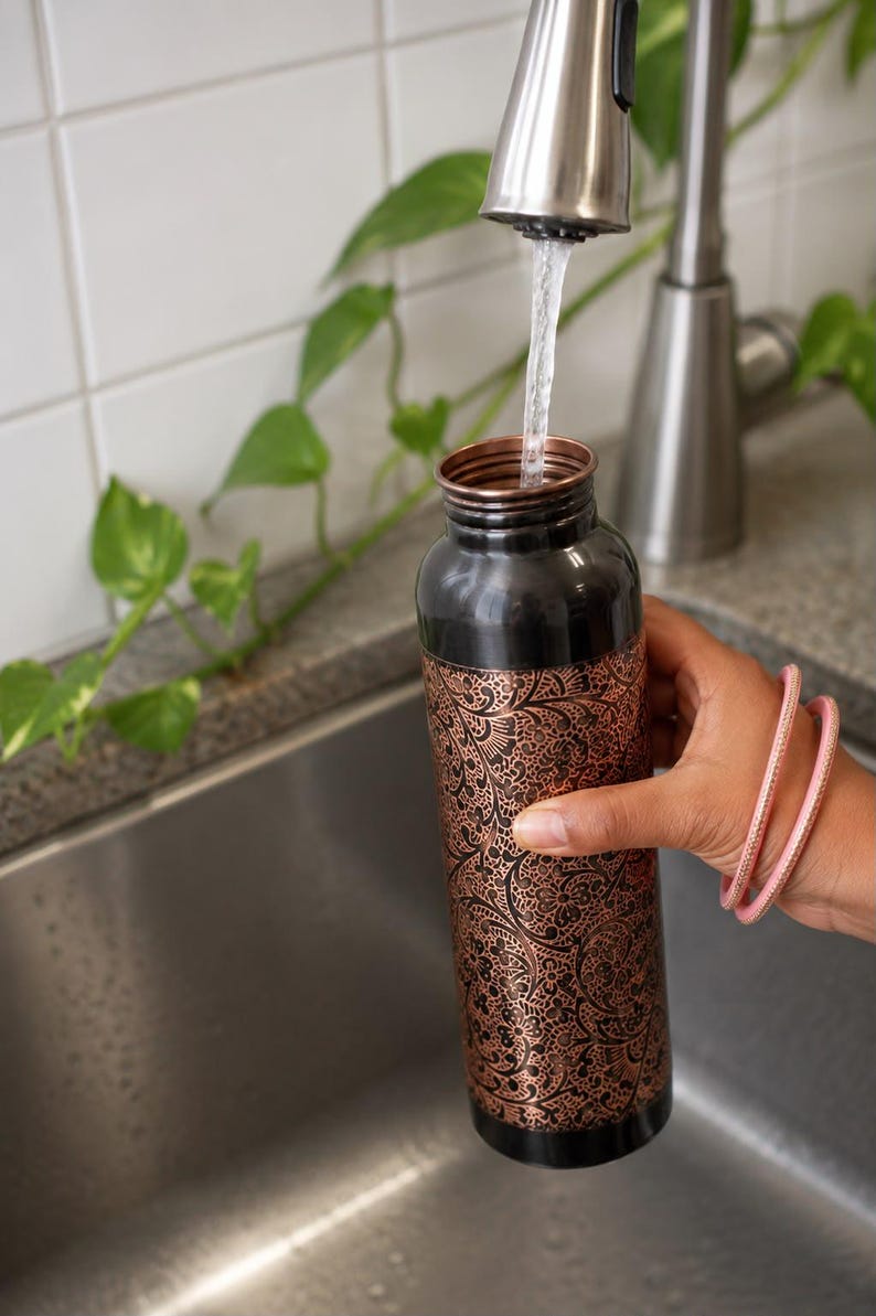K&ouml;nnte beinhalten: Eine zylindrische Wasserflasche aus Kupfer mit schwarzem Finish an Hals und Boden sowie einem detaillierten, ge&auml;tzten Blumenmuster in metallischem Kupfer auf dem Hauptk&ouml;rper. Die Flasche wird gerade unter einem Wasserhahn bef&uuml;llt.