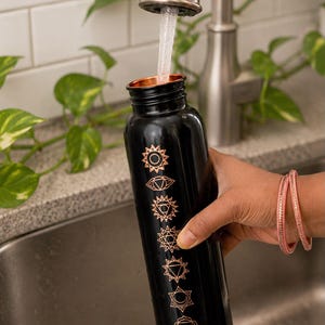 K&ouml;nnte beinhalten: Eine schwarze zylindrische Wasserflasche mit einem kupferfarbenen Metallrand und einer vertikalen Reihe von sieben eingravierten kupferfarbenen Chakrasymbolen. Die Flasche wird mit Wasser aus einem Wasserhahn gef&uuml;llt, wobei ein klarer Strahl.