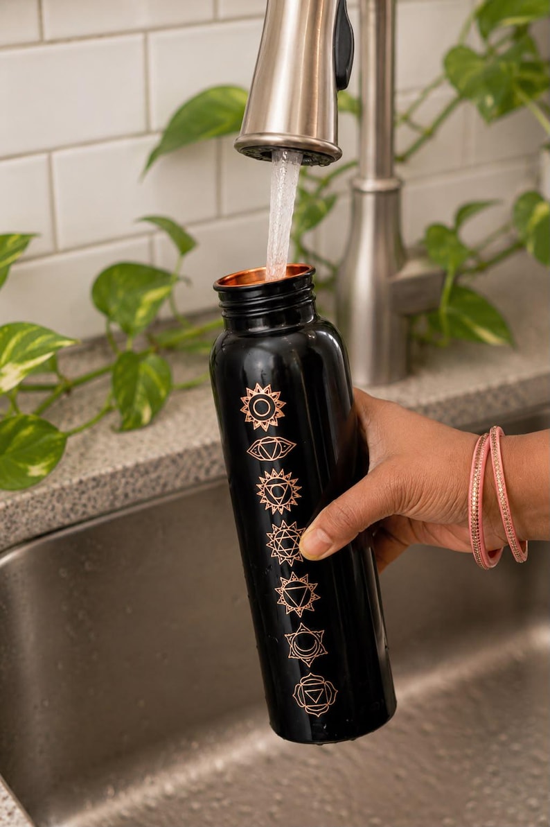 K&ouml;nnte beinhalten: Eine schwarze zylindrische Wasserflasche mit einem kupferfarbenen Metallrand und einer vertikalen Reihe von sieben eingravierten kupferfarbenen Chakrasymbolen. Die Flasche wird mit Wasser aus einem Wasserhahn gef&uuml;llt, wobei ein klarer Strahl.