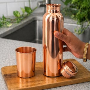 May include: A copper water bottle, cup, and lid sit on a wooden cutting board. The bottle is cylindrical and has a screw-on lid. The cup is short and wide. The items have a shiny, metallic finish. The background includes a kitchen sink and greenery.