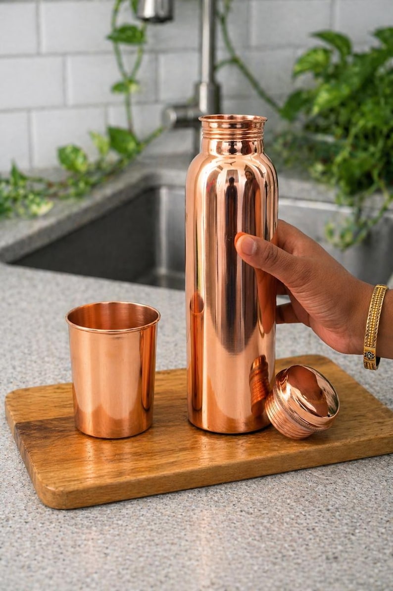 May include: A copper water bottle, cup, and lid sit on a wooden cutting board. The bottle is cylindrical and has a screw-on lid. The cup is short and wide. The items have a shiny, metallic finish. The background includes a kitchen sink and greenery.