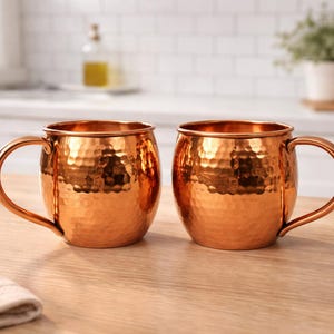 May include: Two copper Moscow Mule mugs with hammered texture and curved handles sit on a wooden surface. The mugs have a shiny, reflective finish. The background includes a kitchen setting with white tile and plants.
