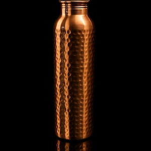Puede incluir: Una botella de agua de color cobre con una superficie texturizada y con hoyuelos. La botella tiene una tapa de rosca y una forma cil&iacute;ndrica. El fondo es negro, resaltando el brillo met&aacute;lico de la botella. La botella mide unos 25 cm de alto.