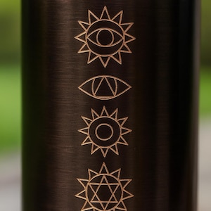 K&ouml;nnte beinhalten: Eine dunkelbraune Metallwasserflasche mit einer vertikalen Reihe aus f&uuml;nf eingravierten Symbolen. Die Motive zeigen ein Auge in einer Sonne, ein dreieckiges Auge, eine kreisf&ouml;rmige Sonne, einen sechszackigen Stern und ein nach unten zeigendes Dreieck.