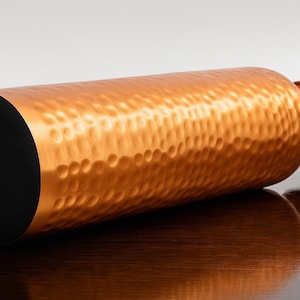 May include: A copper-colored water bottle with a hammered texture. The bottle has a black base and a screw-on cap. The bottle is lying horizontally on a dark surface, reflecting its shape.