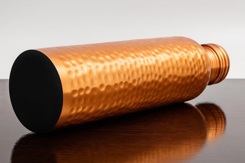 May include: A copper-colored water bottle with a hammered texture. The bottle has a black base and a screw-on cap. The bottle is lying horizontally on a dark surface, reflecting its shape.