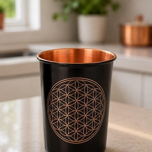 K&ouml;nnte beinhalten: Ein zylindrischer Becher mit einer glatten schwarzen Au&szlig;enseite und einer metallisch kupferfarbenen Innenseite. Auf der Vorderseite befindet sich ein kreisf&ouml;rmiges geometrisches Muster der Blume des Lebens in Kupferfarbe.