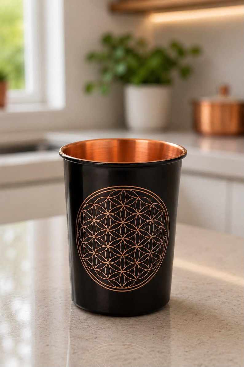 K&ouml;nnte beinhalten: Ein zylindrischer Becher mit einer glatten schwarzen Au&szlig;enseite und einer metallisch kupferfarbenen Innenseite. Auf der Vorderseite befindet sich ein kreisf&ouml;rmiges geometrisches Muster der Blume des Lebens in Kupferfarbe.