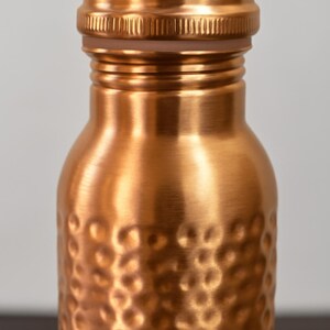 May include: Close-up of a copper-colored water bottle with a textured, hammered finish. The bottle has a screw-on cap and a rounded body. The copper color gives the bottle a warm, metallic appearance. The bottle is designed for reusable use.