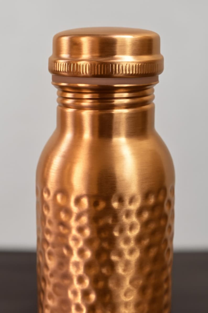 May include: Close-up of a copper-colored water bottle with a textured, hammered finish. The bottle has a screw-on cap and a rounded body. The copper color gives the bottle a warm, metallic appearance. The bottle is designed for reusable use.