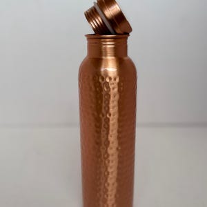 Puede incluir: Una botella de agua de cobre con un acabado martillado texturizado. La botella es cil&iacute;ndrica con una tapa de rosca, parcialmente desenroscada. La botella est&aacute; vac&iacute;a y se encuentra en posici&oacute;n vertical sobre una superficie blanca.