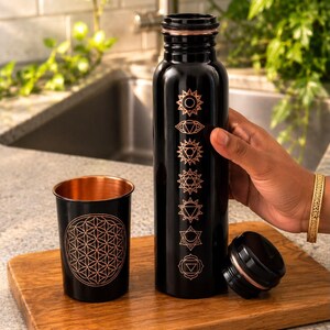 K&ouml;nnte beinhalten: Eine schwarze Metallwasserflasche und ein passender Becher. Die Flasche zeigt sieben vertikal angeordnete kupferfarbene Chakrasymbole. Der Becher ist mit einem kupferfarbenen geometrischen Lebensblumenmuster verziert. Beide haben ein kupfernes Inneres.