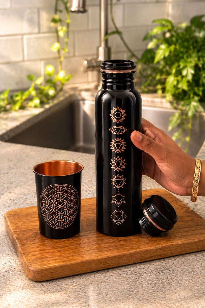 K&ouml;nnte beinhalten: Eine schwarze Metallwasserflasche und ein passender Becher. Die Flasche zeigt sieben vertikal angeordnete kupferfarbene Chakrasymbole. Der Becher ist mit einem kupferfarbenen geometrischen Lebensblumenmuster verziert. Beide haben ein kupfernes Inneres.