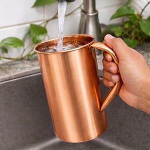 May include: A copper pitcher with a curved handle is held under a faucet as water fills it. The pitcher has a shiny, metallic finish. The background includes a stainless steel faucet, a sink, and green leafy plants.