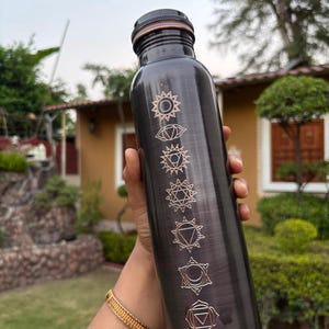 K&ouml;nnte beinhalten: Eine dunkelmetallische Wasserflasche mit geb&uuml;rstetem Finish, die sieben vertikal angeordnete, gravierte Chakrasymbole in einem hellen Kupferton aufweist. Die Flasche hat einen zylindrischen K&ouml;rper und einen passenden dunklen Schraubverschluss.
