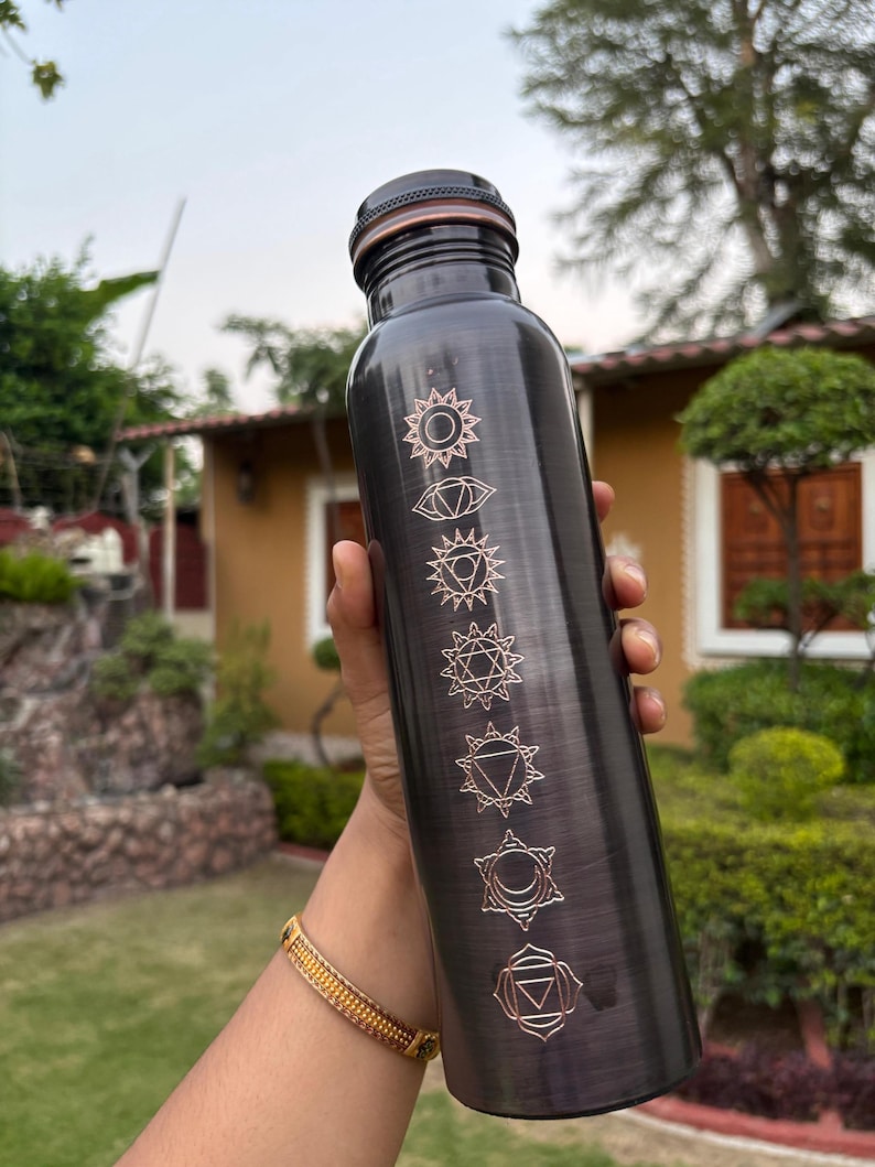 K&ouml;nnte beinhalten: Eine dunkelmetallische Wasserflasche mit geb&uuml;rstetem Finish, die sieben vertikal angeordnete, gravierte Chakrasymbole in einem hellen Kupferton aufweist. Die Flasche hat einen zylindrischen K&ouml;rper und einen passenden dunklen Schraubverschluss.
