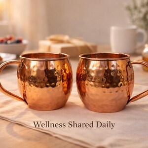 May include: Two copper Moscow Mule mugs with hammered texture and curved handles sit on a white cloth. The mugs are in focus, with a blurred background of a table setting, including a plate of food and the text "Wellness Shared Daily".
