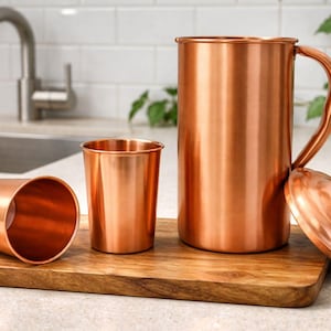 May include: A copper pitcher with a handle, a lid, and two copper cups sit on a wooden cutting board. The pitcher and cups have a shiny, metallic finish. The kitchenware is displayed in a well-lit setting.