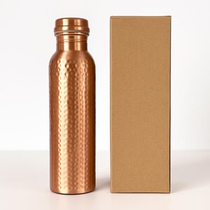 Puede incluir: Una botella de agua de color cobre con textura martillada, junto a una caja de cart&oacute;n marr&oacute;n liso. La botella tiene una tapa de rosca y una forma cil&iacute;ndrica y delgada. La botella mide aproximadamente 23 cm de alto.