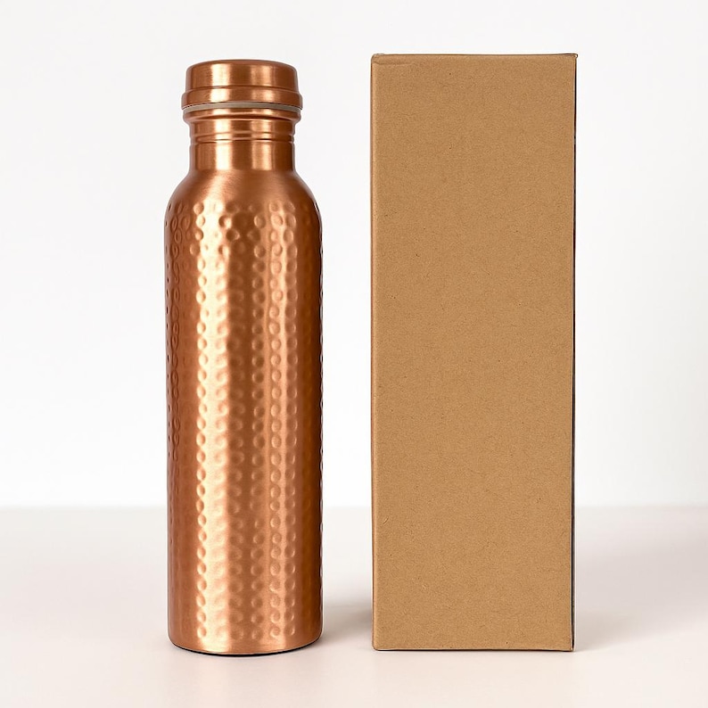 Puede incluir: Una botella de agua de color cobre con textura martillada, junto a una caja de cart&oacute;n marr&oacute;n liso. La botella tiene una tapa de rosca y una forma cil&iacute;ndrica y delgada. La botella mide aproximadamente 23 cm de alto.